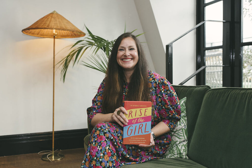 Image of Jo Wimble-Groves on a sofa holding her book, Rise of The Girl